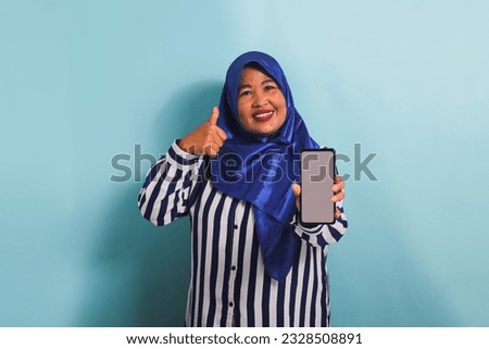 A smiling middle-aged Asian woman in a blue hijab and a striped shirt is gesturing a thumbs-up while holding a phone with a white blank screen. She is isolated on a blue background.