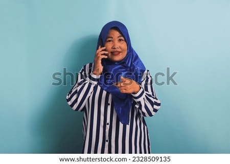 A middle-aged Asian woman in a blue hijab and a striped shirt is pointing at the camera while talking on the phone. She is isolated on a blue background