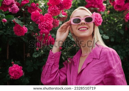 Happy smiling fashionable woman wearing trendy pink retro style sunglasses, linen summer shirt, posing in rose garden, among flowers. Copy, empty space for text Royalty-Free Stock Photo #2328510719