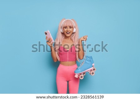 Barbie style woman in pink top and leggins with blue roller-skates, phone and white headphones posing on blue background, vintage fashion concept, copy space
