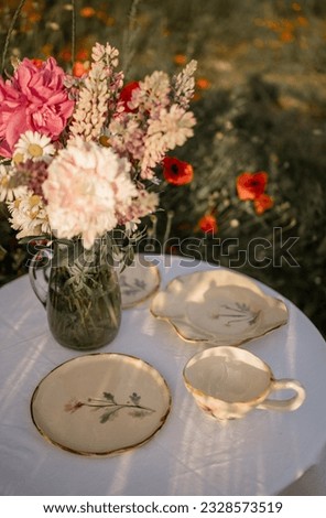 
A table with a white tablecloth, ceramic dishes and a vase of flowers in the middle of a poppy field