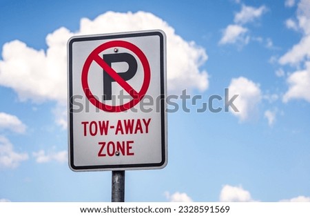 A close up, upwards view of a no parking sign, a black P with a red, crossed out circle. The sign also advises that it is a tow-away zone. There is blue sky and lots of clouds in the background.