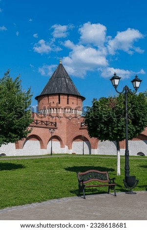 Old Tula Kremlin red brick wall and tower with wooden roof in sunny summer day. Clear blue sky with few clouds. Medieval architecture. Empty bench by green lawn. Travel in Russia theme.