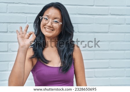 Asian young woman standing over bricks background doing ok sign with fingers, smiling friendly gesturing excellent symbol 