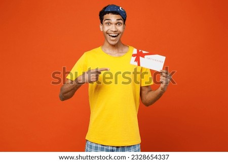 Calm young man wear pyjamas jam sleep eye mask rest relax at home hold point on gift certificate coupon voucher card for store isolated on plain orange background studio. Good mood night nap concept