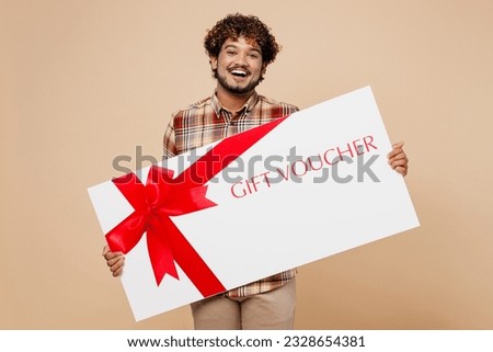 Big young surprised Indian man wearing brown shirt casual clothes hold gift certificate coupon voucher card for store isolated on plain pastel light beige background studio. People lifestyle concept