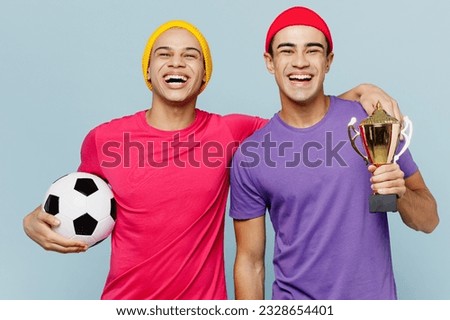 Young fun fans couple two friends men wear casual clothes together cheer up support football sport team hold soccer ball cup watch tv live stream isolated on plain pastel light blue background studio