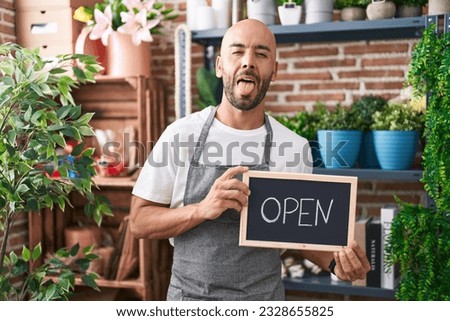 Middle age bald man working at florist holding open sign sticking tongue out happy with funny expression. 