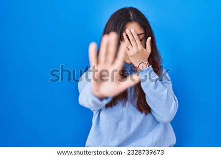 Hispanic young woman standing over blue background covering eyes with hands and doing stop gesture with sad and fear expression. embarrassed and negative concept. 