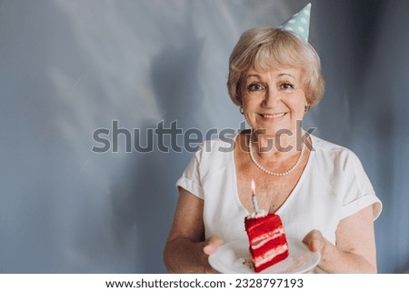 Happy senior woman celebrating her birthday wearing a party hat, holding a cake with a candle on a gray background