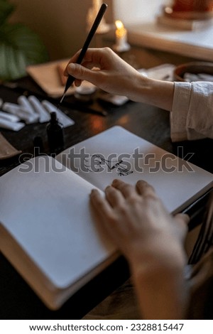 Creative work, passion. Artist sitting at workplace table with burning candles drawing ink in notebook in art studio. Illustrator designer creates new work for freelance order. Hand with fountain pen