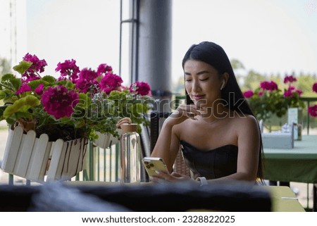 Beautiful young Asian woman sitting at a terrace in a outdoors restaurant and browsing a menu on a mobile phone. Attractive POC female person using a telephone in a cafe