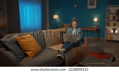 A young woman is lying on the couch in the living room with a laptop on his lap. A woman with a serious face is working or playing on a laptop, watching a movie, photo, video.