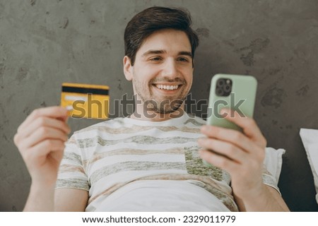Young happy man in casual clothes t-shirt pajama lying in bed use mobile cell phone credit bank card rest relax spend time in bedroom home in own room hotel wake up dream be in reverie good mood day
