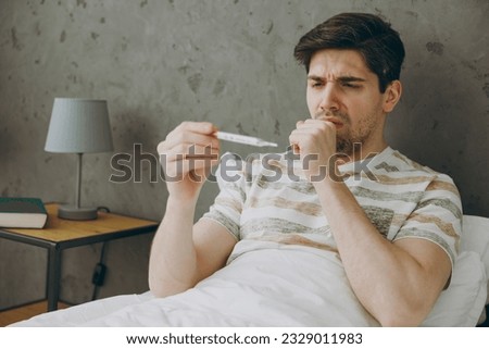 Young sick ill sad man wearing casual clothes t-shirt pajama lying in bed cough measure temperature holding thermometer spend time in bedroom home in own room hotel wake up dream be in bad mood day