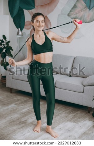 Full body young strong sporty athletic fitness trainer instructor woman wear green tracksuit hold skipping rope behind neck training do exercises at home gym indoor. Workout sport motivation concept
