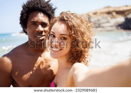 Delighted young woman outstretching hand towards camera while taking selfie with black boyfriend against sea and rocky cliff