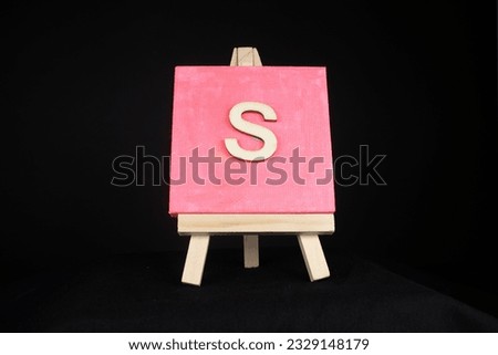 S wooden capital letter and pink blank painting canvas resting on a miniature artists easel isolated on a black background