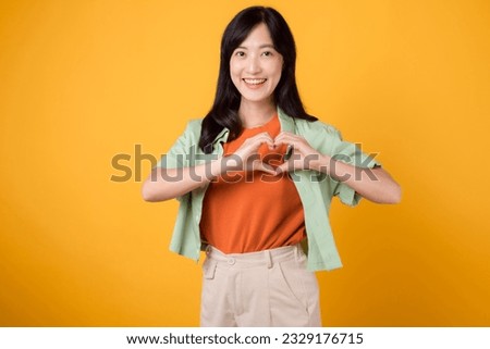 Close up of Asian woman 30s wearing orange shirt showing heart hand gesture, symbolizing warmth and affection. Perfect for expressing love and spreading positive vibes in various projects. Royalty-Free Stock Photo #2329176715