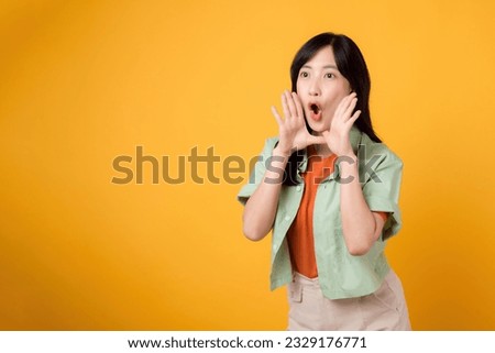Asian cheerful woman 30s wearing orange shirt and green shirt showing hand gesture covering mouth, symbolizing shout or surprise. Energetic moments and conveying sense of exuberance concept.
