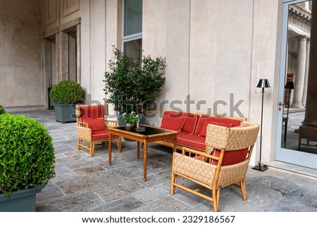 Elegant outdoor lounge made of wicker and red cushions, walnut and glass coffee table, surrounded by plants, in an elegant outdoor loggia.