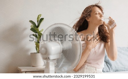 young European woman cools down at home with help of an air conditioner fan and drinks cool water with ice sitting on bed in front of an electric ventilator during intense heat of house, heatstroke Royalty-Free Stock Photo #2329199531