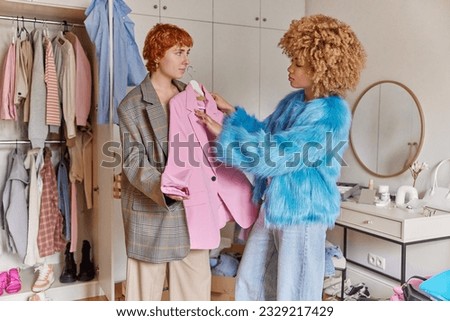 Horizontal shot of two women dressed in fashionable clothes choose what to wear holds pink jacket on hanger poses in bedroom near wardrobe give advice to each other. People and fashion concept