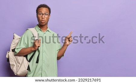 Studio shot of dark skinned man wears spectacles and green shirt points index finger at empty space for your promotional content isolated over purple backgrund. People and lifestyle concept.
