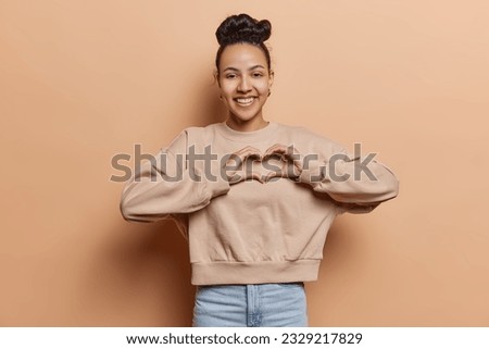 Studio shot of pretty young Latin woman makes heart gesture over chest expresses love and care dressed in casual pullover and jeans isolated over brown background. People and romantic feelings