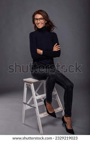 Attractive brunette haired woman sitting against isolated grey background. Brunette haired mid aged woman wearing black sweater and pants. Full length shot. Copy space.