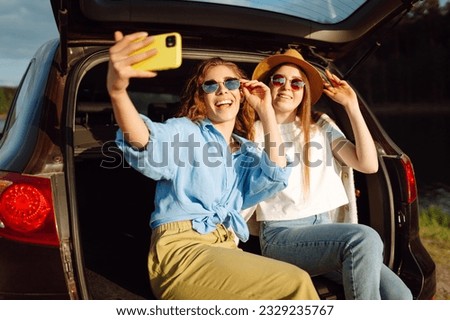Smiling young woman have fun in the back seat of a car while traveling. Girlfriends take selfies, laugh and relax while sitting in car and enjoying the sunny weather. Active lifestyle.