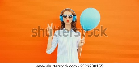 Portrait of happy cheerful young woman in wireless headphones listening to music with balloon on orange background