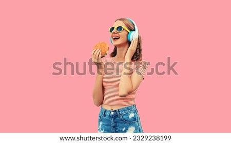 Portrait of happy laughing young woman listening to music in headphones with burger fast food on pink background