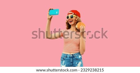 Portrait of modern cheerful laughing young woman taking selfie with phone and burger fast food on pink studio background