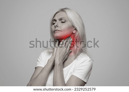 Sick mature lady having sore throat, touching her neck with red inflamed zone, suffering from laryngeal disorder, tonsillitis, throat cancer, cold, posing on studio background, black and white photo Royalty-Free Stock Photo #2329252579