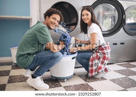 Happy Young Multicultural Couple Doing Laundry Together Loading Washer Machine At Laundromat Room. Guy Washing His Clothes And Posing Smiling At Camera. Public Laundrette Service Royalty-Free Stock Photo #2329252807