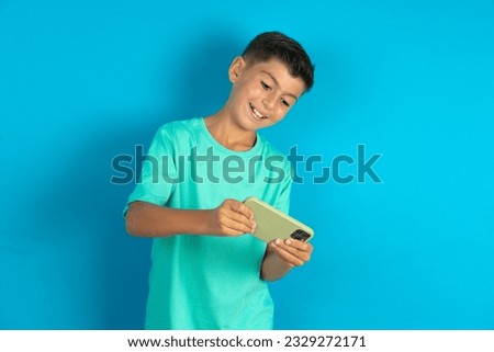 Little hispanic kid boy wearing green T-shirt holding in hands cell playing video games or chatting