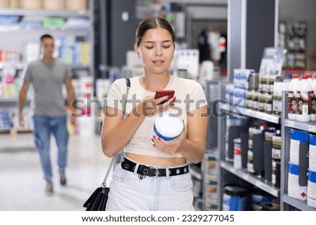 Female customer takes pictures with mobile phone camera of can of paint in department of paints and varnishes. Blurry male buyer in background