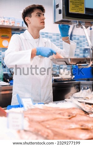 Proficient young salesman in white work clothes weighing scaldfish on scales in fish store with large assortment