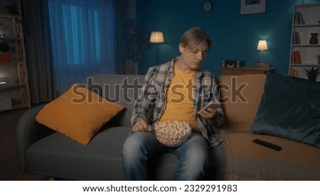 A man is sitting on the couch with a plate of popcorn in front of the TV. A man not interested in a movie sits with a phone in his hands, typing a message, checking email, social network. Boring film.