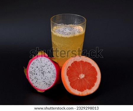 Freshly squeezed tropical fruit juice with seeds in a tall faceted glass on a black background, next to half a ripe pitaya and grapefruit. Close-up.