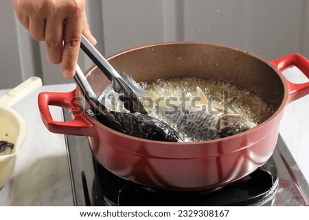 Female Hand using Stainless Tongs, Process of Frying River Carp Fish in a Red Frying Pan. Goreng Ikan Mas  Royalty-Free Stock Photo #2329308167
