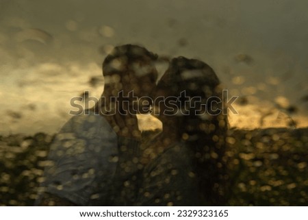 blurred image of a young couple guy and girl kissing on the lips at sunset in the rain, shot through glass with raindrops. High quality photo Royalty-Free Stock Photo #2329323165