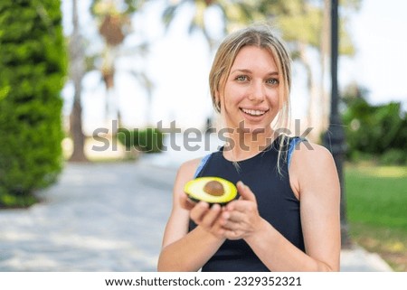 Young blonde woman at outdoors holding an avocado with happy expression