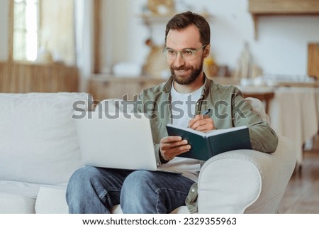 Handsome smiling bearded businessman wearing eyeglasses using laptop computer, taking notes, watching training courses at home. Smart student studying, learning language, online education concept 
