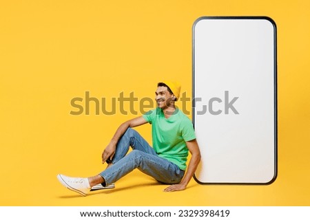 Full body smiling happy fun young man he wearing casual clothes green t-shirt hat sitting near big huge blank screen mobile cell phone smartphone with area isolated on plain yellow background studio