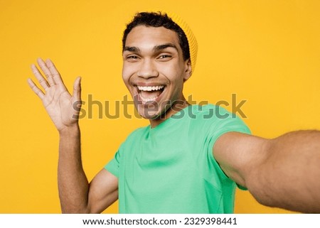 Close up young man of African American ethnicity wears casual clothes green t-shirt hat doing selfie shot pov on mobile cell phone waving hand isolated on plain yellow background. Lifestyle concept