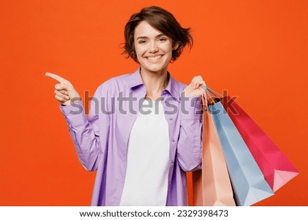 Young happy woman wear purple shirt casual clothes hold in hand paper package bags after shopping point aside on area isolated on plain orange color background studio Black Friday sale buy day concept
