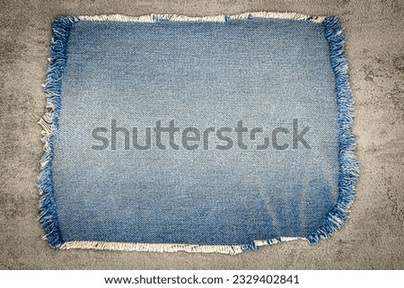 Denim blue jeans fabric frame on grey cement background. Ripped denim cloth swatch. Destroyed torn denim blue jeans patch. Recycle old jeans denim piece. Sample fragment of jeans cloth. Royalty-Free Stock Photo #2329402841