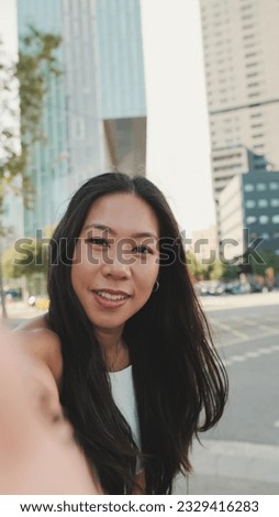 Close up, cute happy girl dressed in white top and jeans makes selfie on mobile phone while standing on modern city background